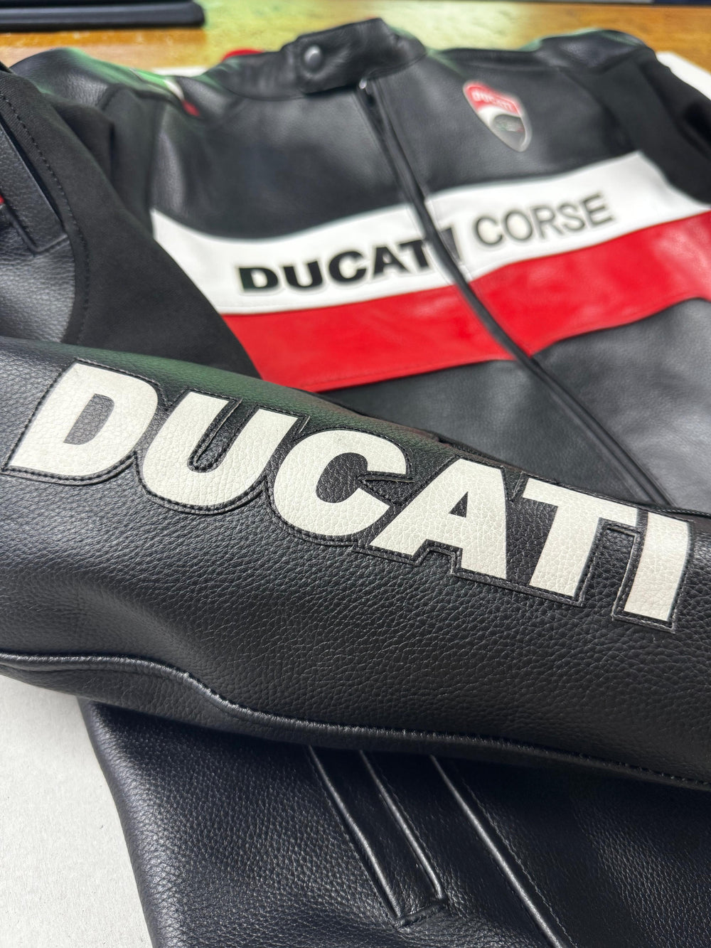 Close-up of the Ducati Corse C5 Men's Motorcycle Leather Jacket, focusing on the 'Ducati' branding on the sleeve and the 'Ducati Corse' logo on the chest. The image highlights the high-quality black leather with red and white accents, showcasing the jacket's premium craftsmanship.