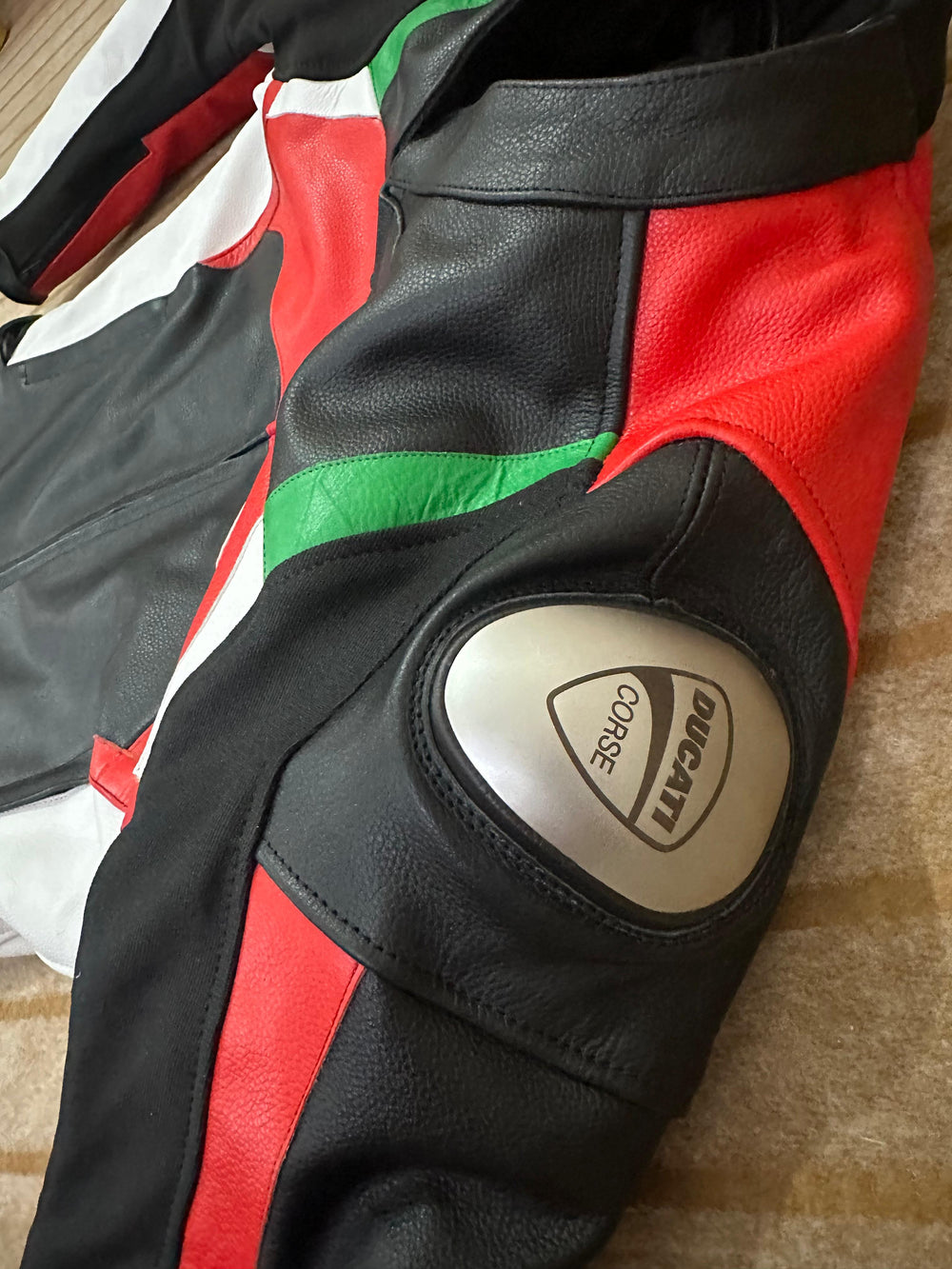 Close-up view of the Ducati Corse C3 Leather Motorbike Jacket, focusing on the shoulder area with protective armor. The image highlights the detailed texture of the black, red, and green leather, showcasing the jacket's durable construction and stylish design